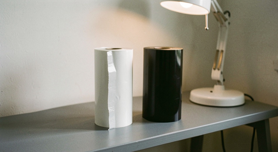 Two rolls of vinyl film standing upright on a grey bench under a desk lamp, the cut edge of one visibly thicker and whiter than the other